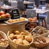 Nahaufnahme eines Teils des Frühstücksbuffets im Best Western Hotel Achim Bremen mit mehreren Körben voller frischer Brötchen und Brot. Daneben steht eine Schale mit Äpfeln und anderen Früchten sowie Gebäck. Im unscharfen Hintergrund sind Tische und Stühl