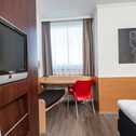 A cozy single room features a wall-mounted TV, a modern red chair at a small desk, and abstract art above the bed.