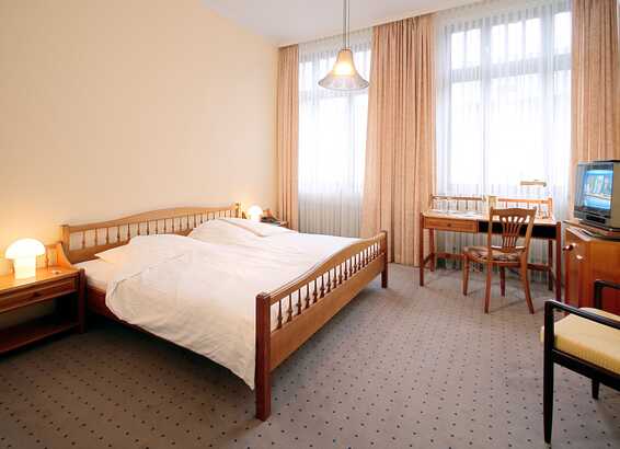 A cozy double room features a wooden bed, a vintage TV, and a desk by large windows with beige curtains.