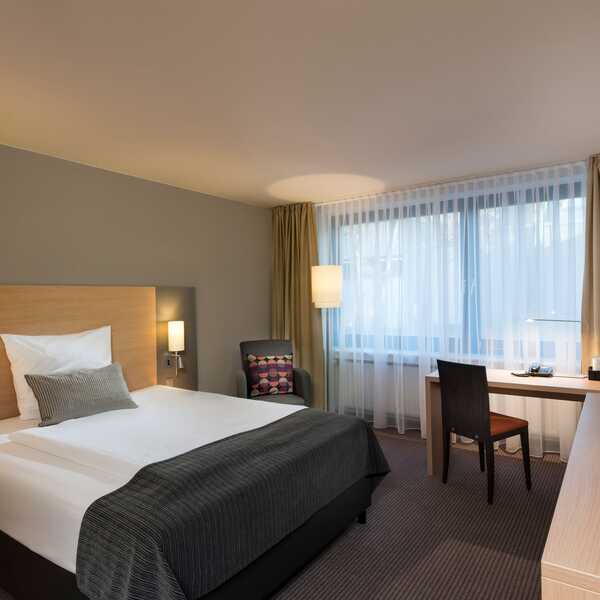 Ein modernes Einzelzimmer mit gemütlichem Bett, Schreibtisch und großem Fenster bietet eine entspannte Standardatmosphäre.