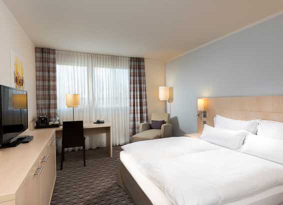 A standard hotel room features a cozy bed, a TV, and a desk by the window with sheer curtains.