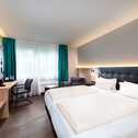 Ein modernes und komfortables Doppelzimmer im Best Western Hotel Achim Bremen. Es verfügt über zwei Einzelbetten mit dunklem Kopfteil und weißer Bettwäsche. Links vom Bett befindet sich ein Schreibtisch mit Stuhl und einem Fernseher an der Wand. Der Raum