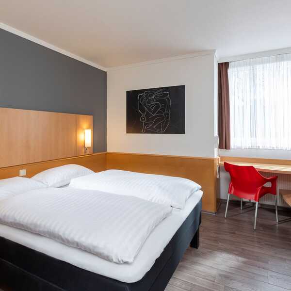 Ein modernes Standardzimmer mit einem Doppelbett, einem Schreibtisch und einem roten Stuhl unter einem großen Fenster.