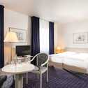 A cozy double room features twin beds, a small table with chairs, and a TV, all set against elegant dark curtains.