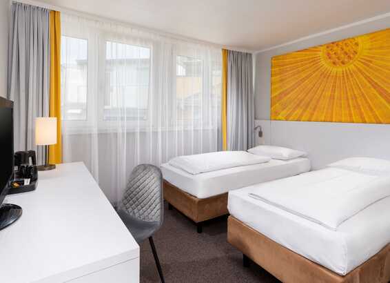 Zwei Einzelbetten stehen in einem hellen Zimmer mit gelbem Wandbild, großem Fenster und einem Schreibtisch mit Fernseher.