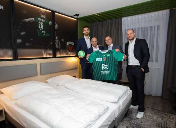 Four men in suits hold a green sports jersey and a ball in a hotel room, showcasing team spirit.