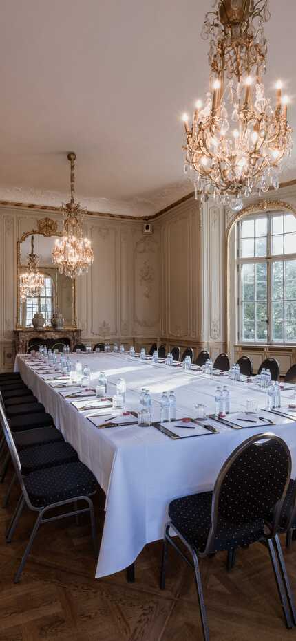 A grand music room features an elegant conference table with chairs, under ornate chandeliers and large windows.