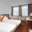 A twin bed room features two neatly made beds, a desk with a computer, and large windows with beige curtains.
