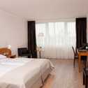 A spacious hotel room features a neatly made bed, a desk with a coffee maker, and large windows with elegant curtains.