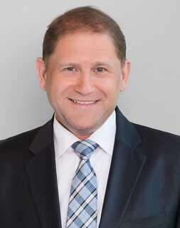 A man in a suit and tie smiles confidently against a plain background.