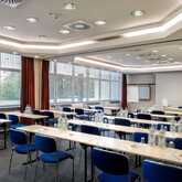 A spacious meeting room with rows of tables and blue chairs is set up with water bottles and notepads, ready for a conference.