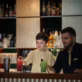 A bartender smiles as a young boy eagerly reaches for a colorful drink at a bar counter.