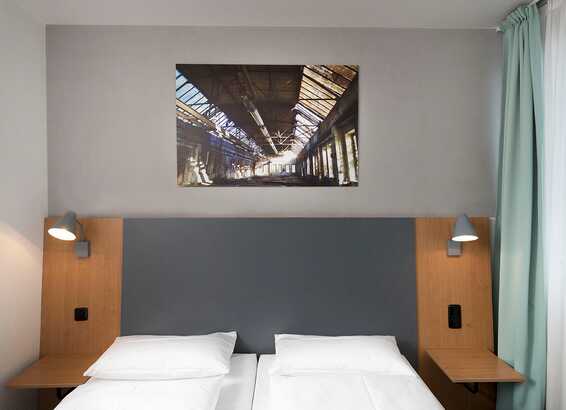 A minimalist double room features twin beds, modern lighting, and a large industrial-themed artwork above the headboard.