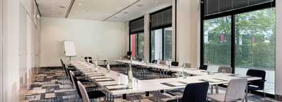 A modern meeting room with a U-shaped table setup, chairs, and a whiteboard, overlooking a garden through large windows.