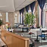 A stylish restaurant interior features elegant table settings with blue accents and lush plants against large windows.