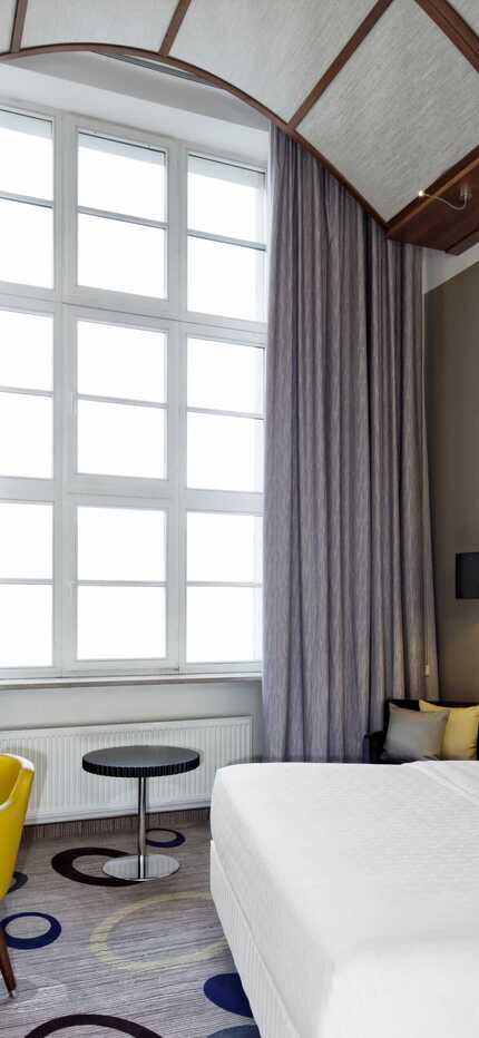 A modern hotel room features a large window, cozy bed, and stylish decor with a yellow chair and elegant lighting.