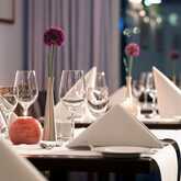 Elegant restaurant tables are set with folded napkins, wine glasses, and purple flowers in sleek vases.