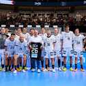 A handball team in white jerseys poses with a coach holding a commemorative jersey on a vibrant indoor court.