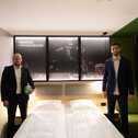 Two men in suits stand beside a hotel bed, one holding a handball, under a sign reading "Hier ist unser Revier".