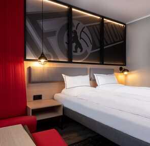 A modern hotel room features a cozy double bed, stylish red accents, and warm lighting, creating a welcoming atmosphere.