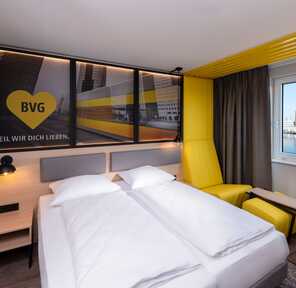 A modern hotel room features a large bed, vibrant yellow accents, and a window view of a riverside cityscape.