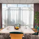 A large, bright conference room in the Best Western Hotel Achim Bremen, prepared for a meeting. A long rectangular table stands in the centre, surrounded by modern chairs with teal-green upholstery. On the table are bottles of water, cups, notepads and a