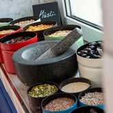 Assorted breakfast ingredients, including seeds and dried fruits, are displayed in colorful bowls with a mortar and pestle nearby.