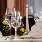 A banquet table is elegantly set with folded napkins, wine glasses, and a floral centerpiece, illuminated by a single lit candle.