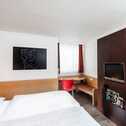 A modern room features a neatly made bed, abstract wall art, a TV, and a cozy workspace with a red chair by the window.