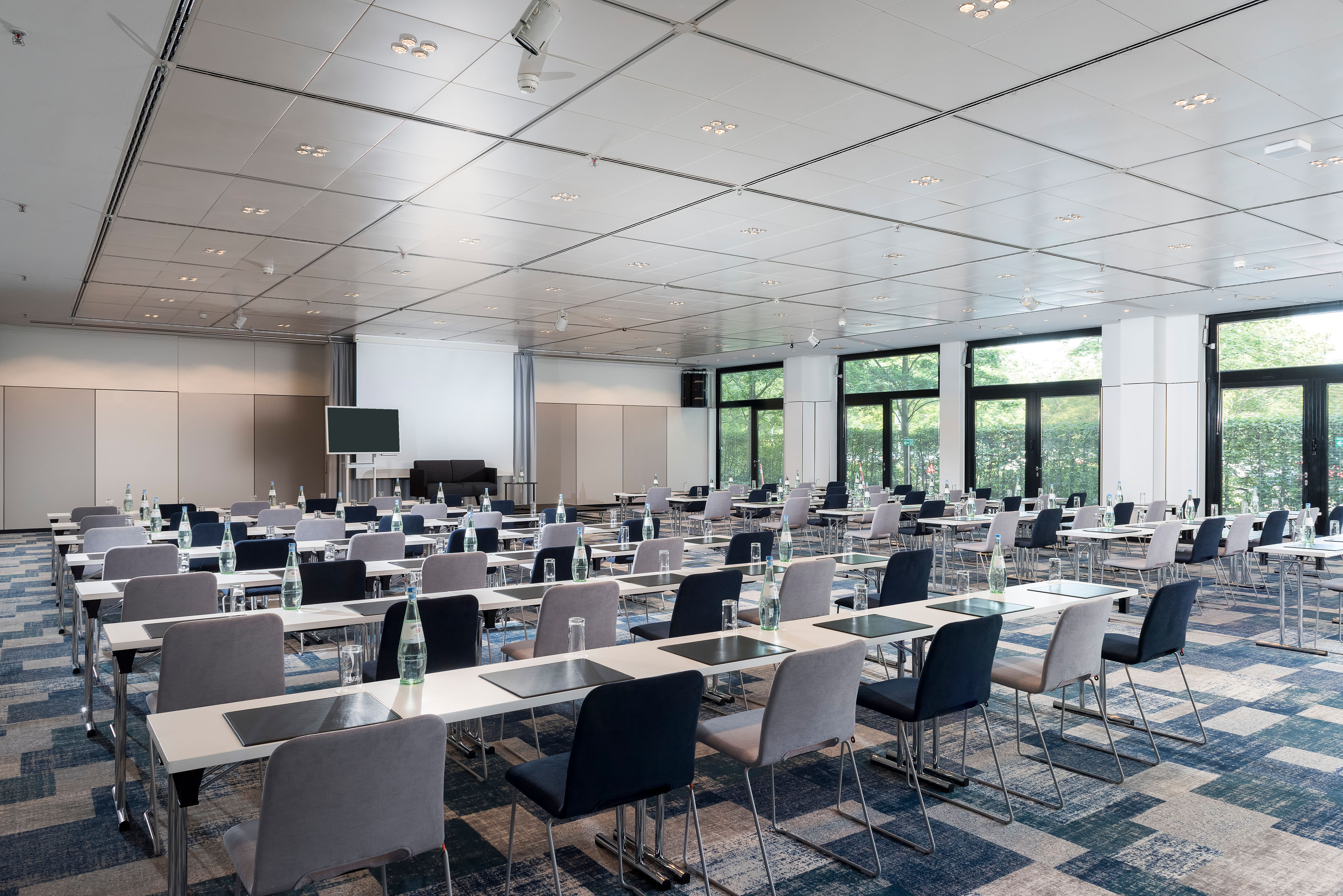 Ein moderner Konferenzraum mit vielen Tischen und Stühlen ist für ein Meeting vorbereitet, mit einem Bildschirm vorne.